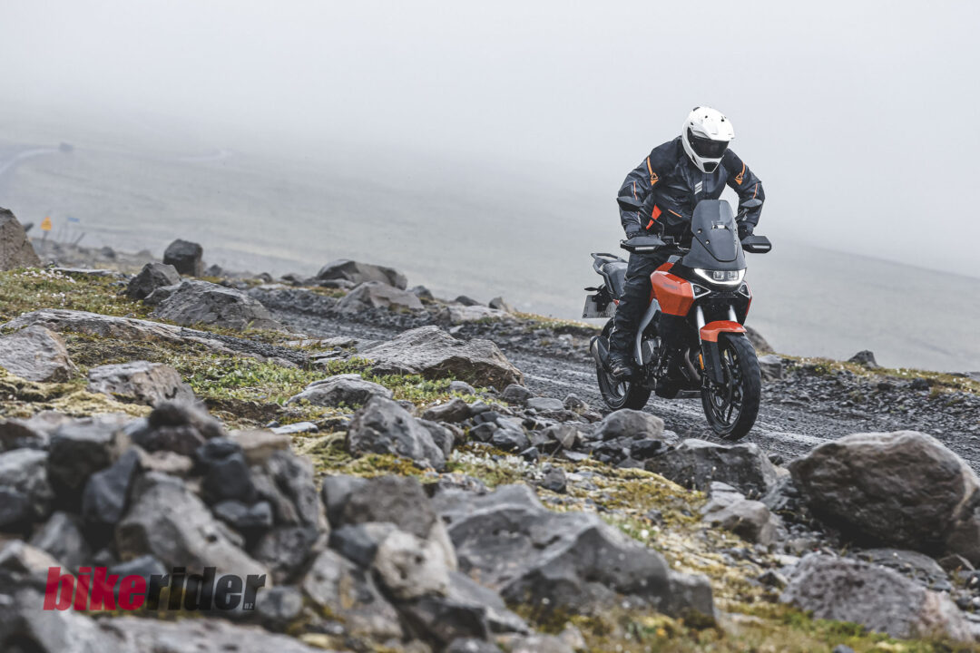 Motorcyclist in white helmet and protective gear rides a red adventure bike along a rocky coastal trail in foggy weather