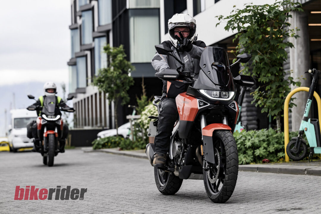 Two motorcyclists riding on a city street; the front rider wears a white helmet and points toward the camera with a gloved hand, as another rider follows behind.