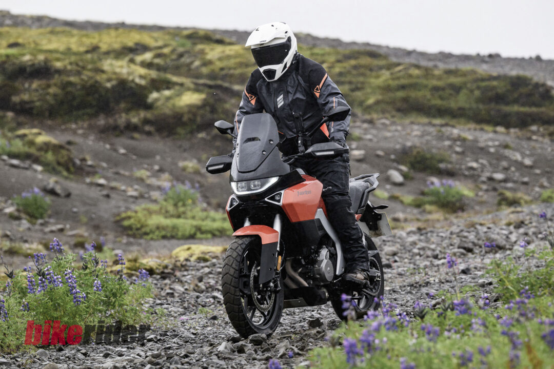 20260712-Norton_Atlas_2026_BC_Paul-Lance-2961-13-1080x720 Motorcyclist in full protective gear riding a red adventure bike on a rocky trail with purple flowers in the foreground