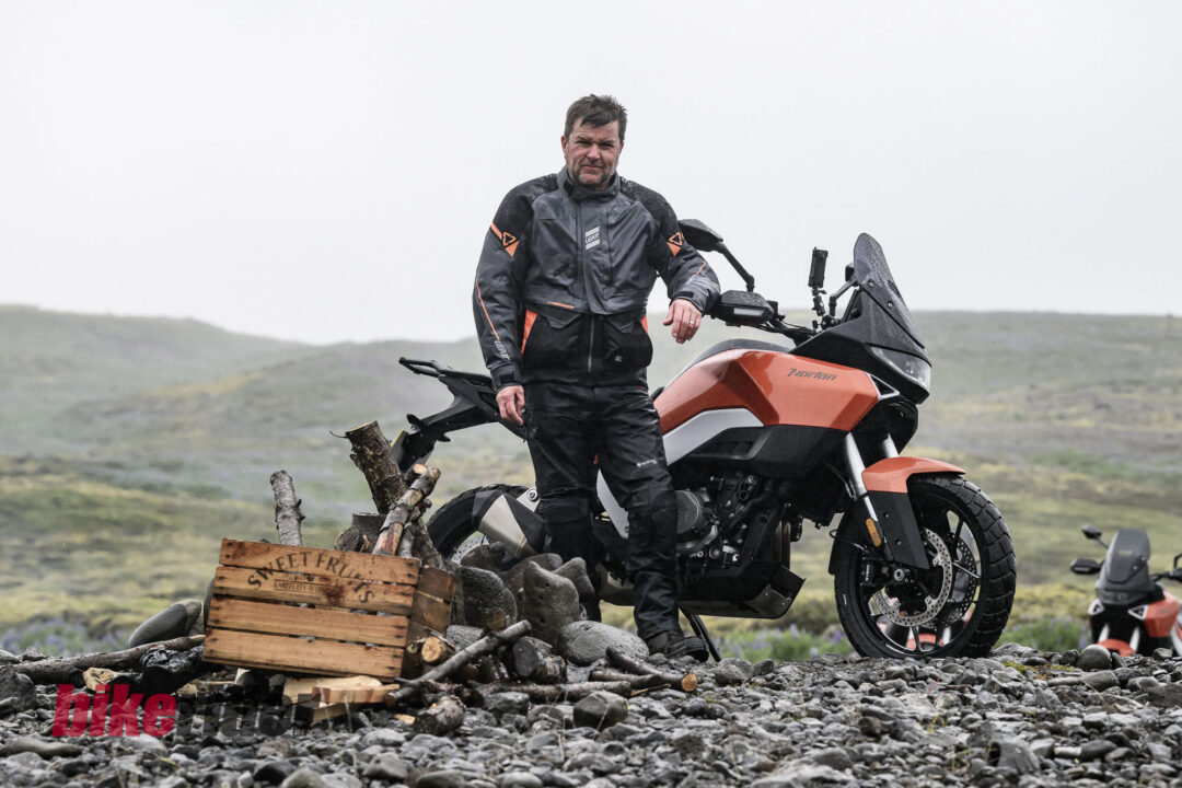 20260712-Norton_Atlas_2026_BC_Paul-Lance-3036-14-1080x720 Man in Leatt protective motorcycle gear stands beside an orange adventure motorcycle on a rocky shoreline, with logs and debris nearby.