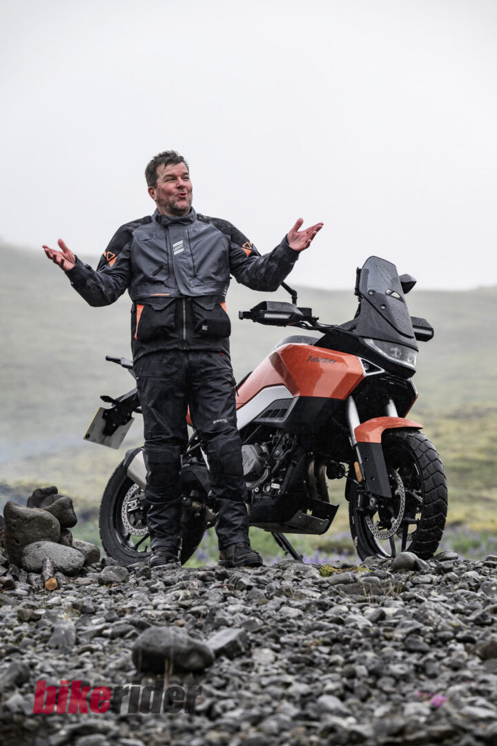 20260712-Norton_Atlas_2026_BC_Paul-Lance-3056-2-720x1080 Man in full motorcycle gear stands with arms outstretched beside an orange adventure motorcycle on a rocky coastal terrain, cloudy sky above.