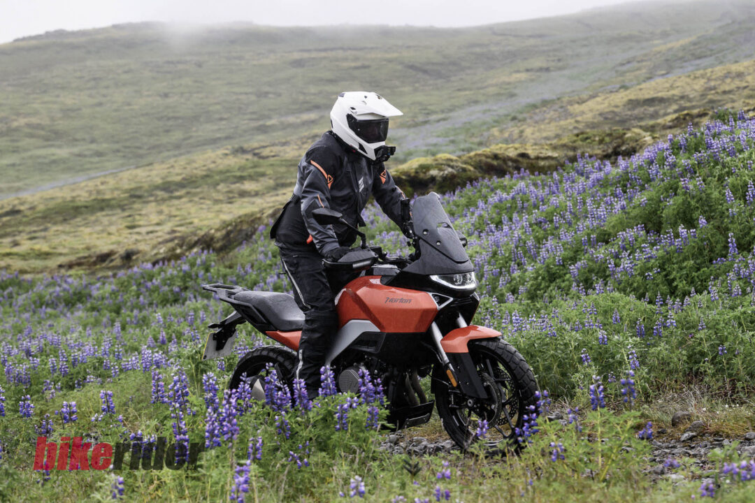 Motorcyclist in full gear and white helmet riding an orange adventure bike through a field of purple wildflowers on a hilly landscape