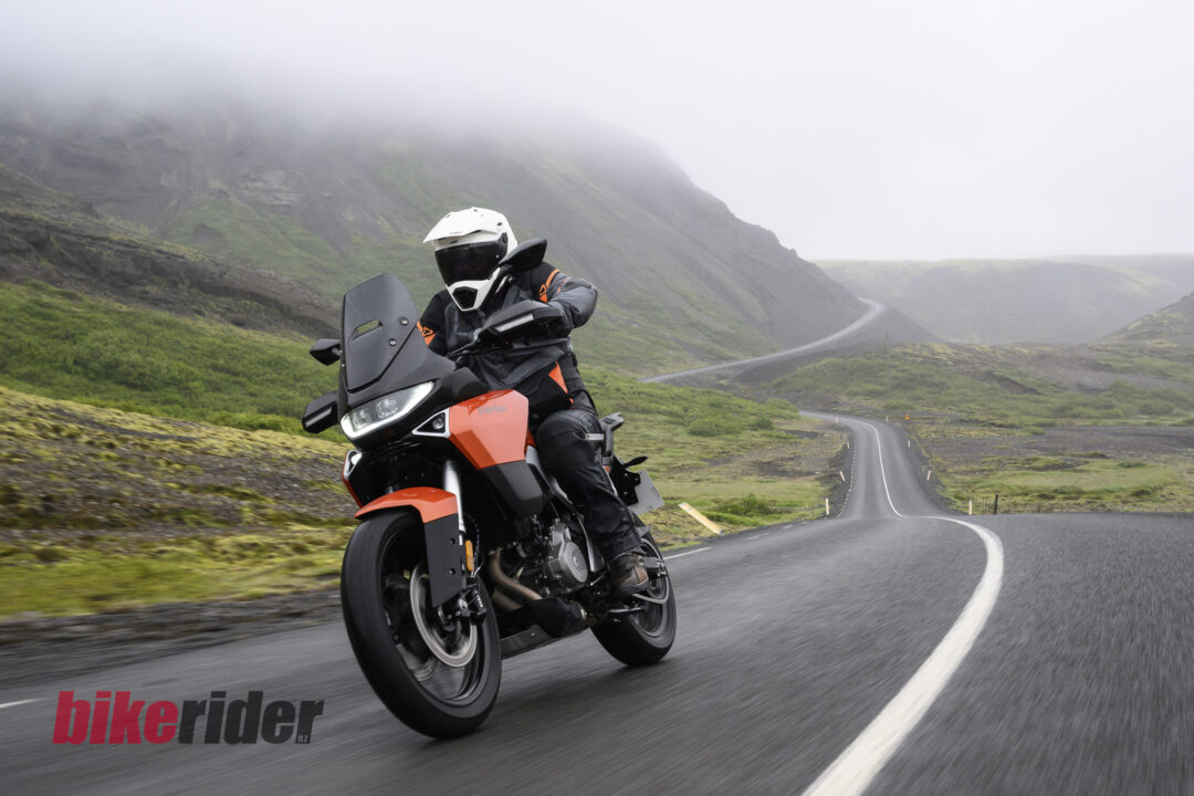 Motorcyclist in a white helmet and black protective gear rides an orange adventure bike on a winding, misty mountain road with green slopes in the background.