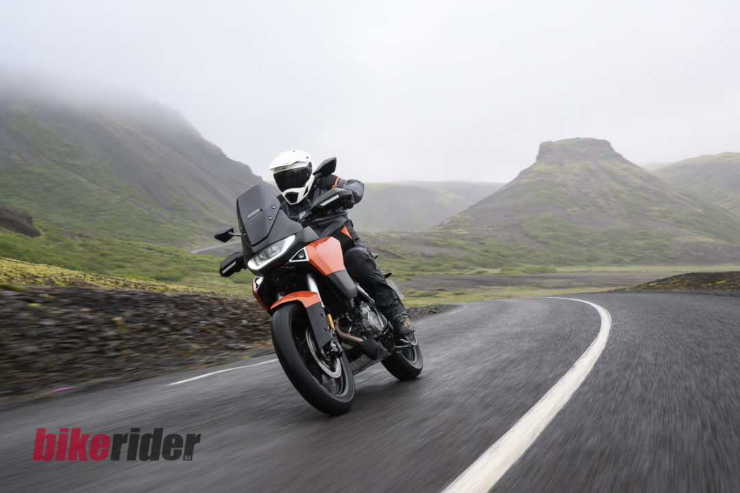 Motorcyclist riding a Norton Atlas adventure bike in white Leatt helmet leaning into a sharp curve on a misty winding mountain road with green hills in the background
