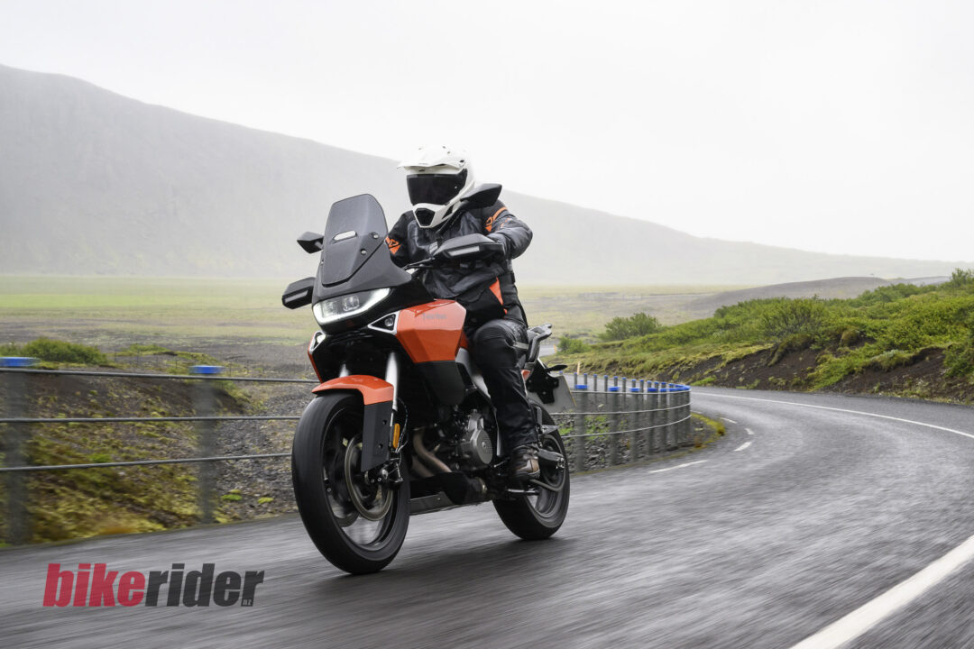 Motorcycle rider in full protective gear on an orange adventure bike navigating a sharp bend on a coastal road; watermark bottom left reads 'bikerider'