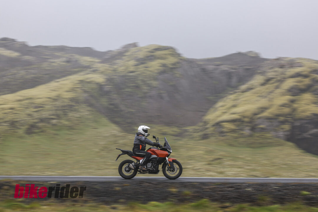 20260715-Norton-Atlas-Global_AB__6925-8-1080x720 Motorcyclist wearing a helmet and dark protective gear rides a red motorcycle along a road with blurred grassy hills in the background.
