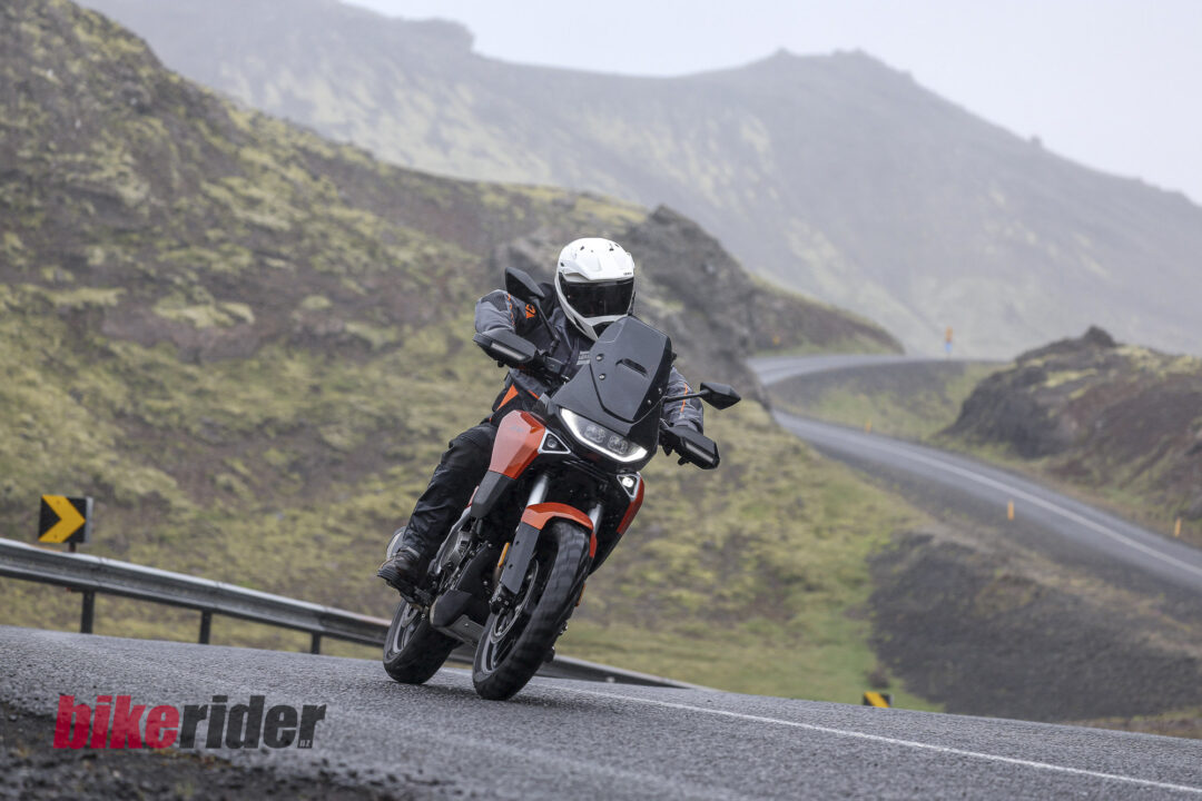 20260715-Norton-Atlas-Global_AB__6955-9-1080x720 Motorcyclist in full gear leaning into a left-hand bend on a winding mountain road, white helmet and orange bike with guardrails nearby.