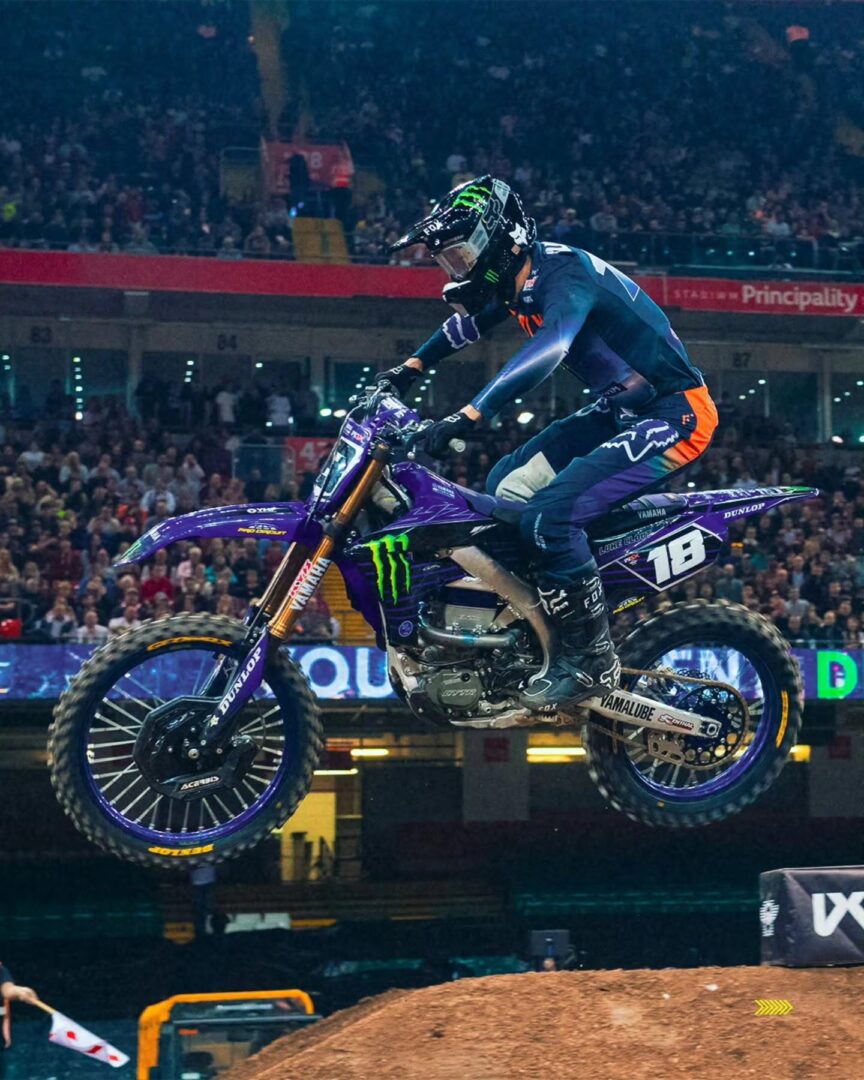 Motocross rider in blue gear mid-air on a purple Yamaha bike during a stadium race, filled with spectators in the background.