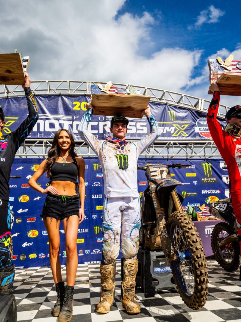 Motocross podium celebration with riders holding wooden trophies above their heads and a smiling presenter beside them on a checkered stage.