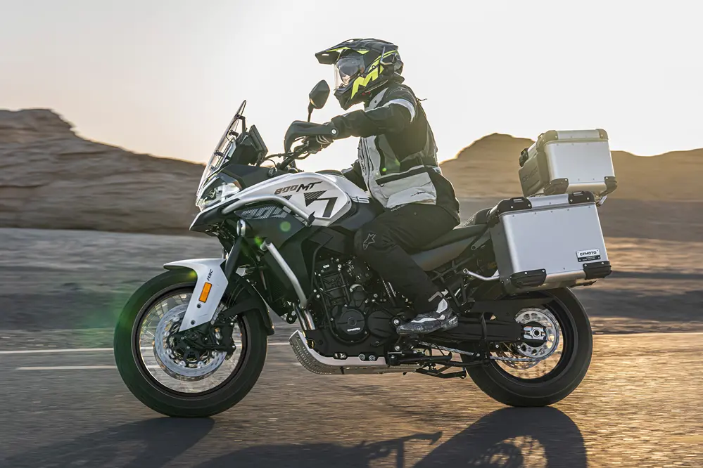 Motorcyclist wearing a helmet and protective gear rides a white adventure motorcycle with silver luggage cases along a desert road at sunset.
