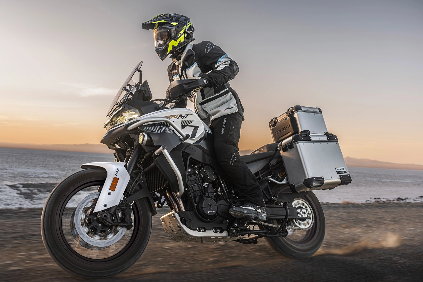 Motorcyclist in full gear riding a silver adventure bike with side and top cases along a coastal road at sunset
