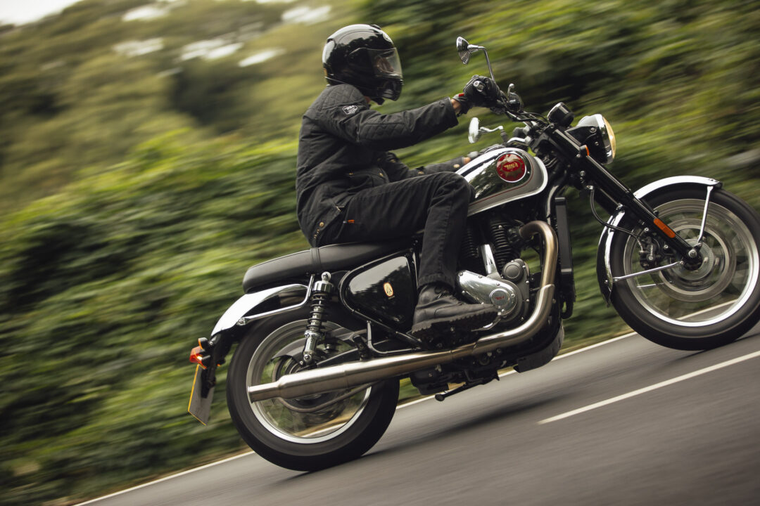 Motorcyclist in black protective gear and helmet riding a black motorcycle at speed on a blurred road surrounded by greenery.