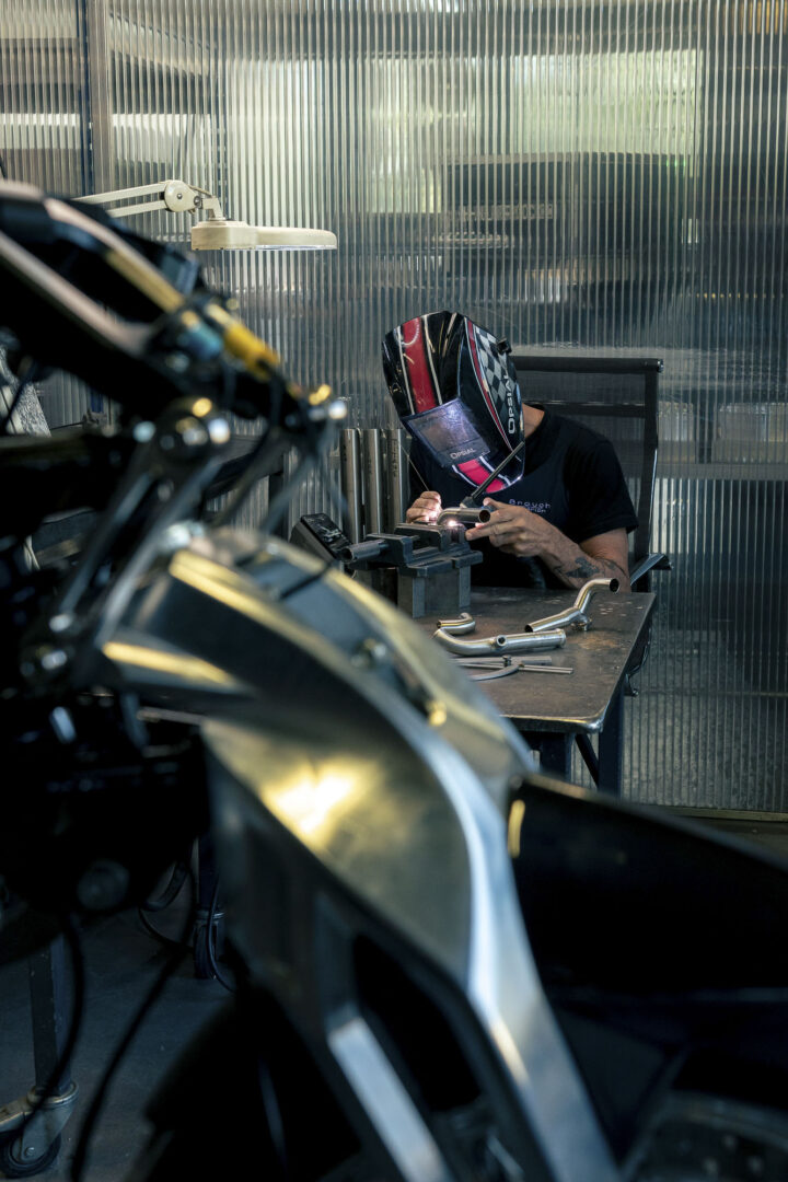 Brough-superior-rmb-01-1-1 Welder in a protective helmet at a workbench, welding a metal piece in a workshop with motorcycle parts nearby.