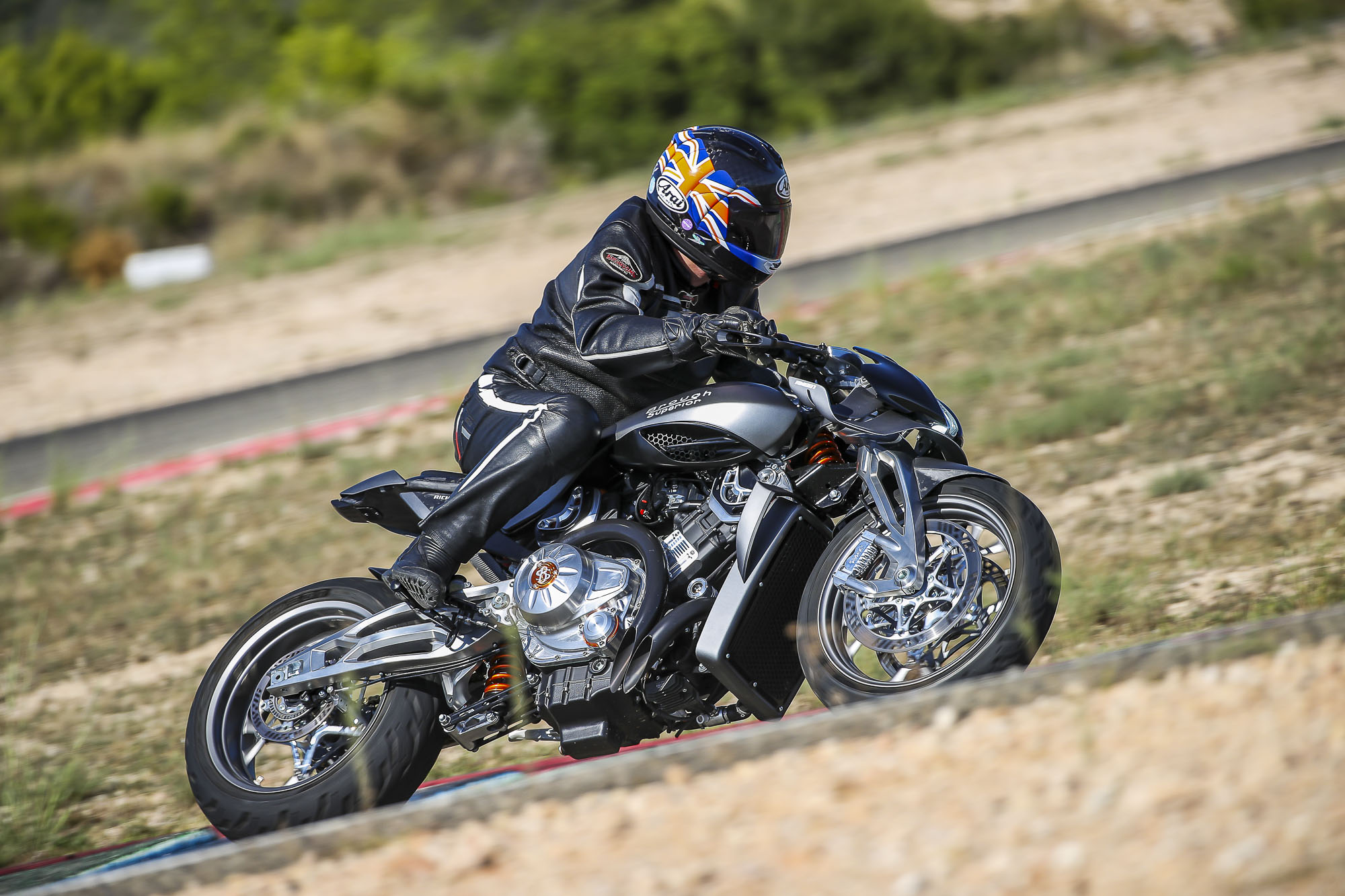 Brough-superior-rmb-01-1 Motorcyclist in black racing suit leans into a turn on a sport bike on a track, helmet featuring a Union Jack design.