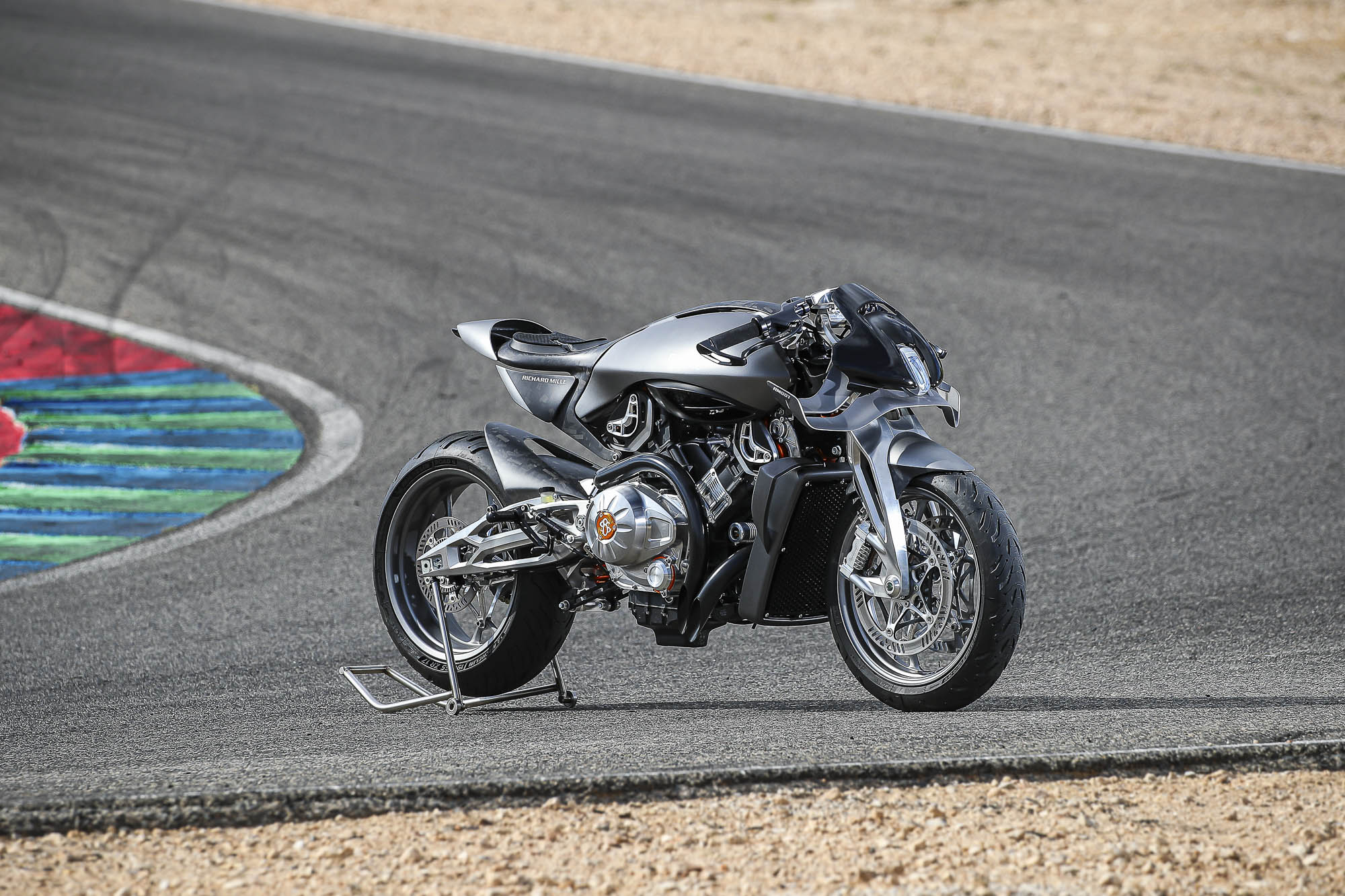 Brough-superior-rmb-01-10-2 Silver sport motorcycle on a race track with a rear stand supporting the wheel, at a corner edge.