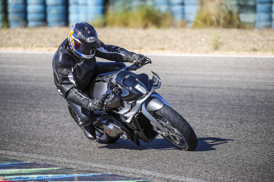 Motorcyclist in black protective gear leaning into a turn on a sportbike on a race track with blue barriers in the background.