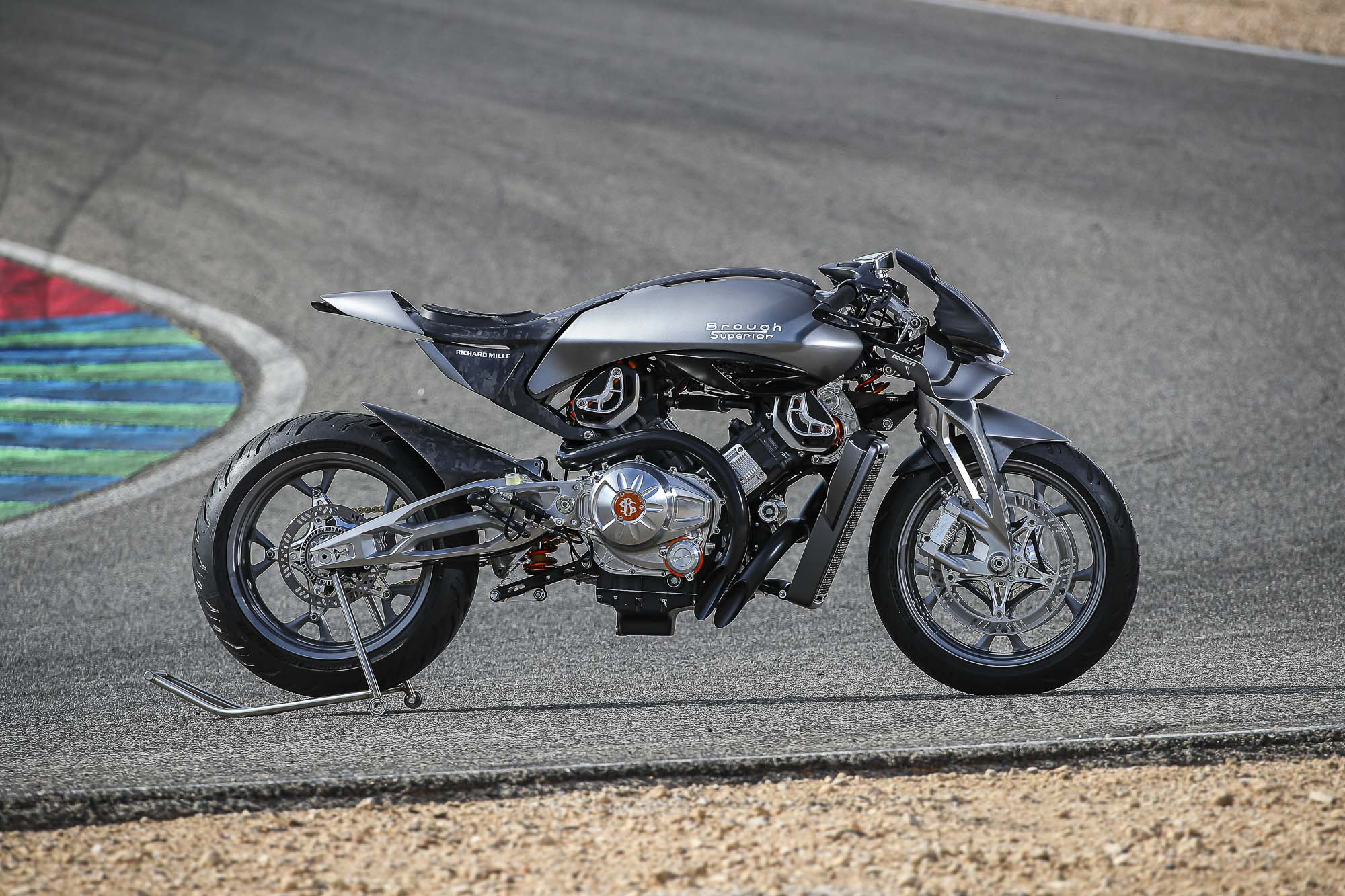 Brough-superior-rmb-01-11-2 Silver Brough Superior motorcycle on a race track, propped on a metal stand with a colorful curb to the left.