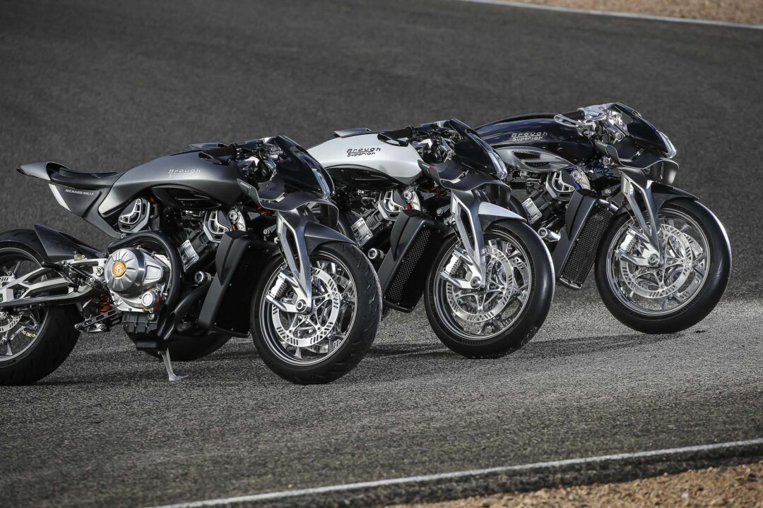Brough-superior-rmb-01-12-2 Row of three shiny motorcycles parked on a racetrack, showing chrome engines and silver/black finishes.