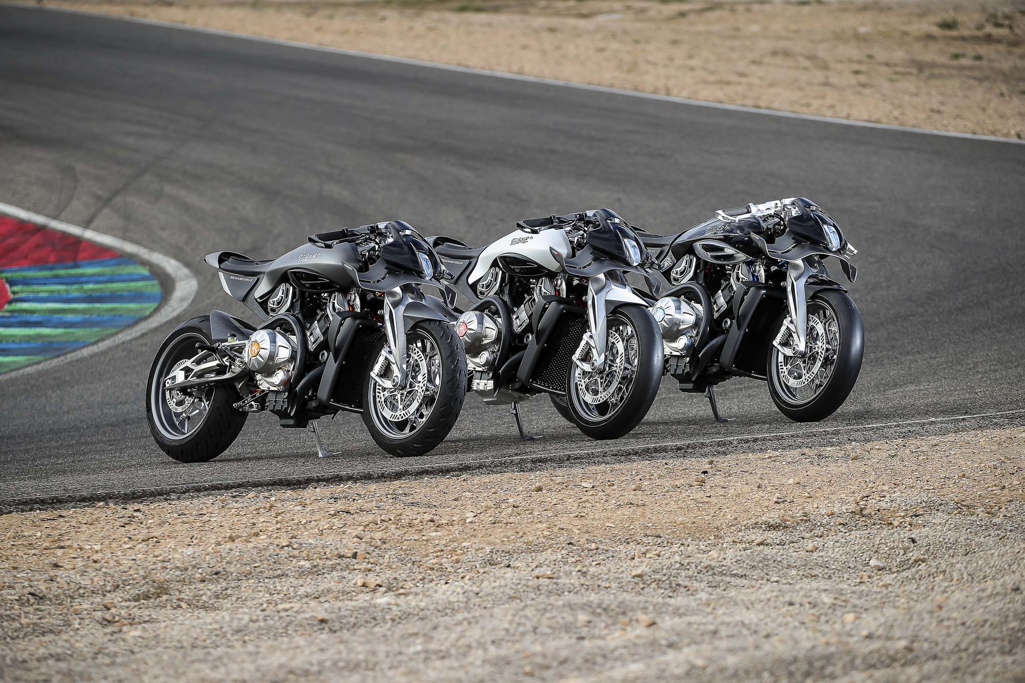 Brough-superior-rmb-01-13-1 Line of silver sport motorcycles leaning into a right-hand turn on a race track on a sunny day.