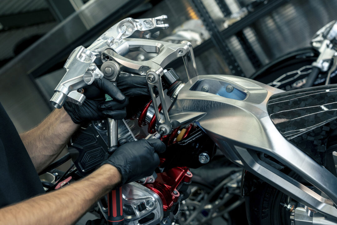 Brough-superior-rmb-01-14 Hands in black gloves assembling a silver motorcycle frame with precision tools in a workshop.
