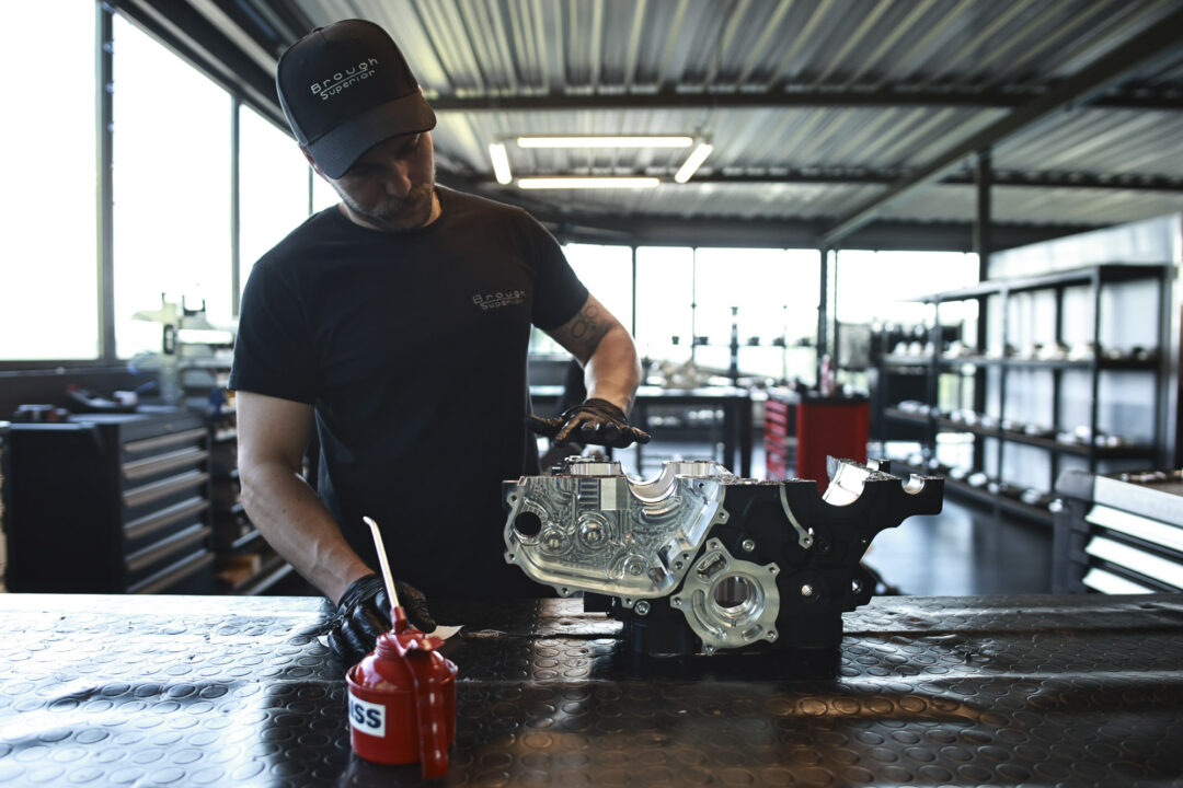 Brough-superior-rmb-01-19 Technician in a black shirt and cap works on a metal engine block at a well-lit workshop, with a red oil can in the foreground.