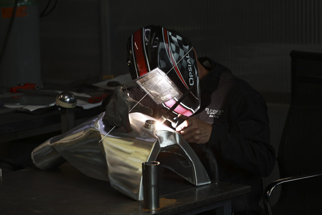 Brough-superior-rmb-01-20 Welder in a dark workshop wearing a red‑black helmet, welding a metal sheet with a bright arc.