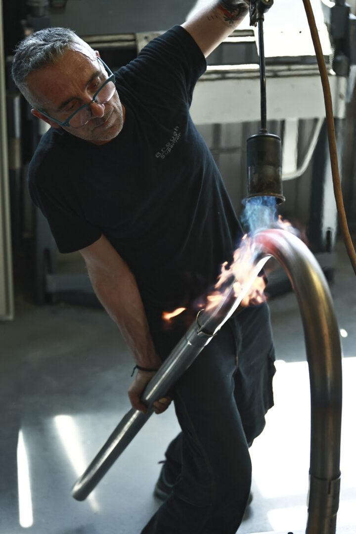 Brough-superior-rmb-01-21 Man in safety glasses and a black shirt holds a heated metal rod with a blowtorch producing blue flames in a workshop.