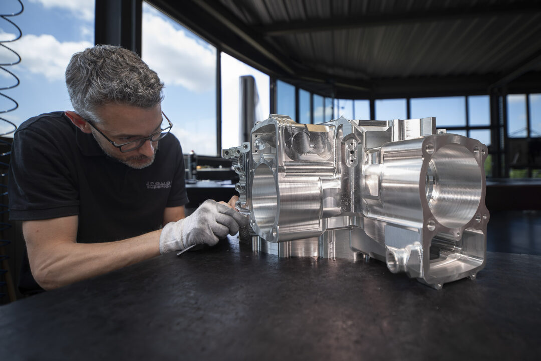 Male technician wearing glasses and gloves inspecting a large machined engine block in a bright workshop.
