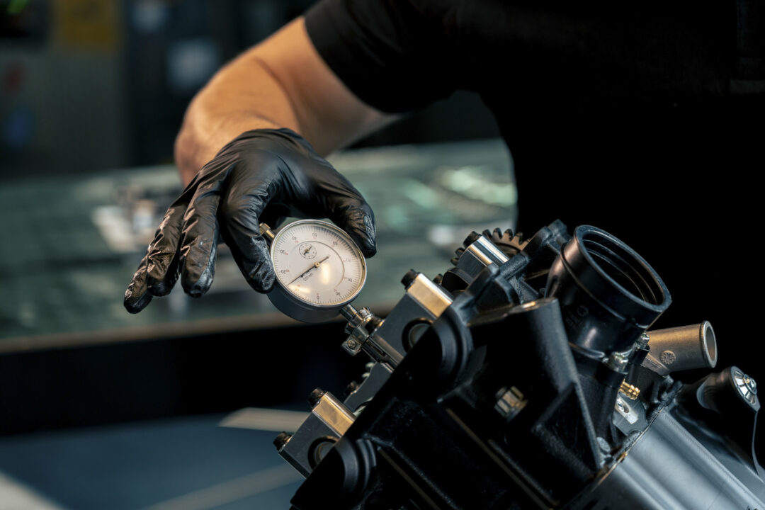 Brough-superior-rmb-01-3-1 Gloved hand holding a dial gauge against a metal mechanical assembly for precision measurement.