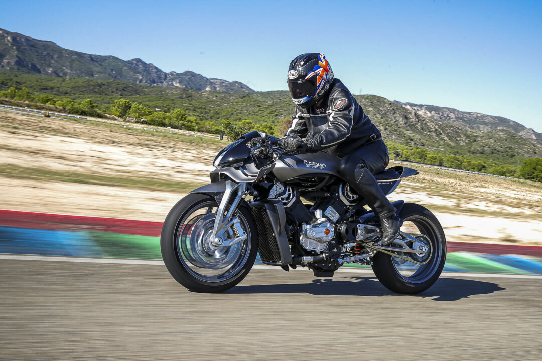 Motorcyclist in black riding gear on a sports bike racing along a track with motion blur foreground and mountains in background.