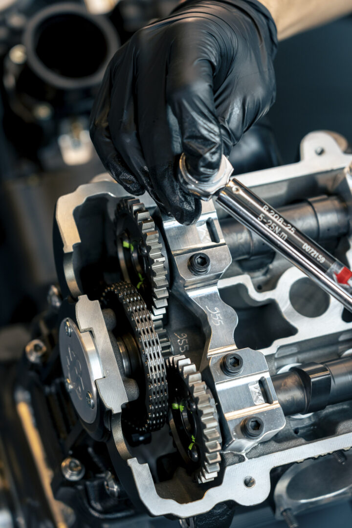 Brough-superior-rmb-01-4-1 Gloved hand using a wrench to tighten gears inside a metallic engine assembly (close-up).