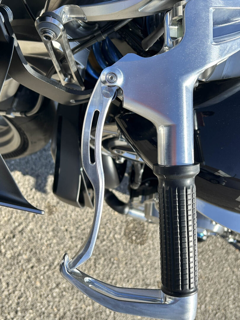 Brough-superior-rmb-01-4-2 Close-up of a chrome motorcycle footpeg and front suspension with a blue frame and black tire in the background.