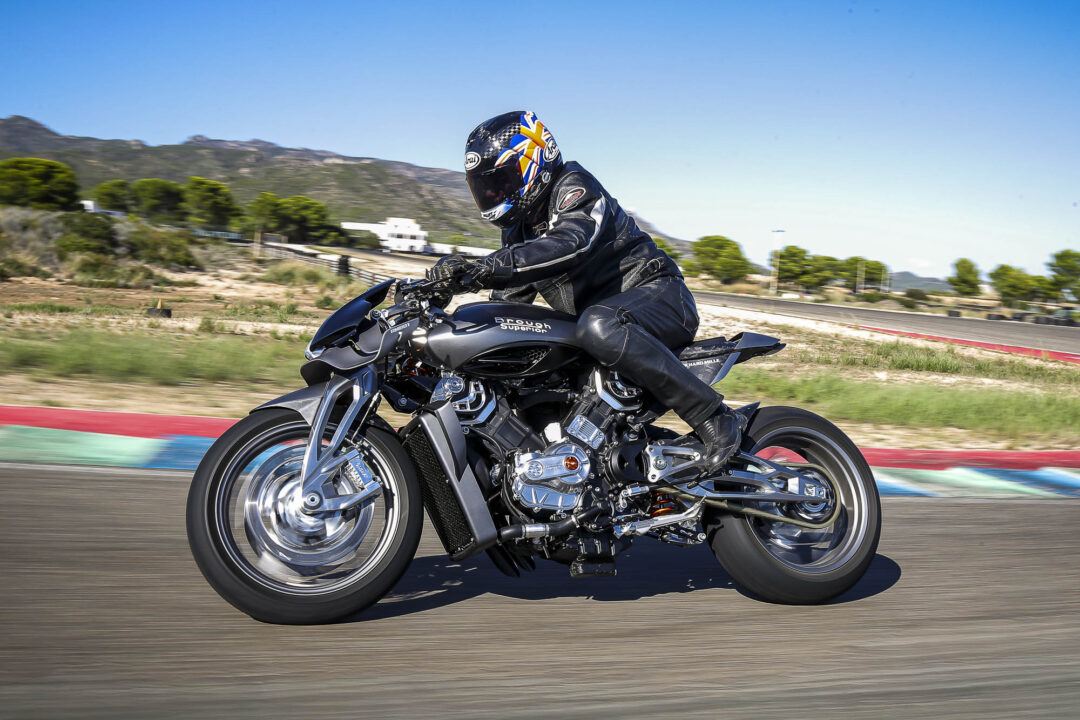 Brough-superior-rmb-01-5 Motorcyclist in a black race suit and colorful helmet speeds along a racetrack, leaning into a turn with motion blur behind him.
