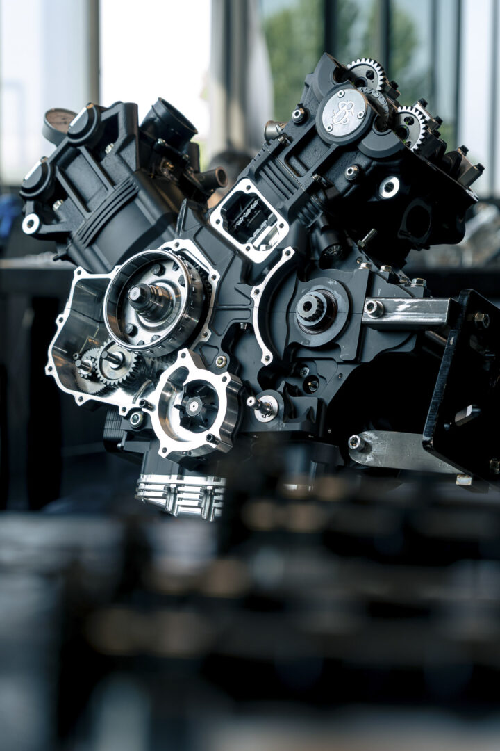 Brough-superior-rmb-01-6-1 Close-up of a black industrial engine block with gears and metal housings in a machining setting.