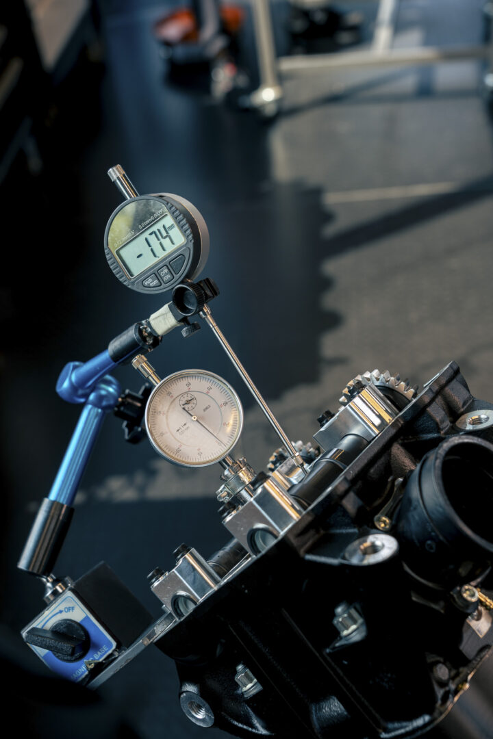 Brough-superior-rmb-01-7-1 Close-up of a hydraulic test rig showing a digital pressure gauge and an analog gauge attached to metal fittings and blue tubing.