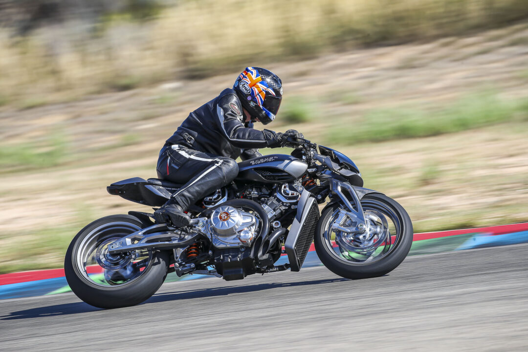 Brough-superior-rmb-01-7 Rider on a blue sport motorcycle leaning into a curve on a racetrack, wearing a black leather suit and a colorful Union Jack helmet.