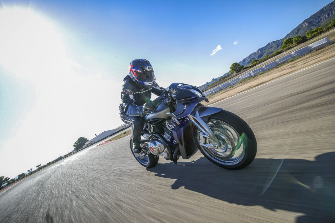 Brough-superior-rmb-01-8 Motorcyclist leaned into a turn on a blue sport bike on a sunny road with mountains in the background; rider wears a full-face helmet with a Union Jack design.
