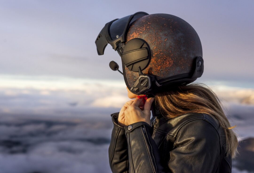 Person wearing a rust-speckled helmet with a built-in headset, adjusting the red strap under a leather jacket on a mountain at sunset.