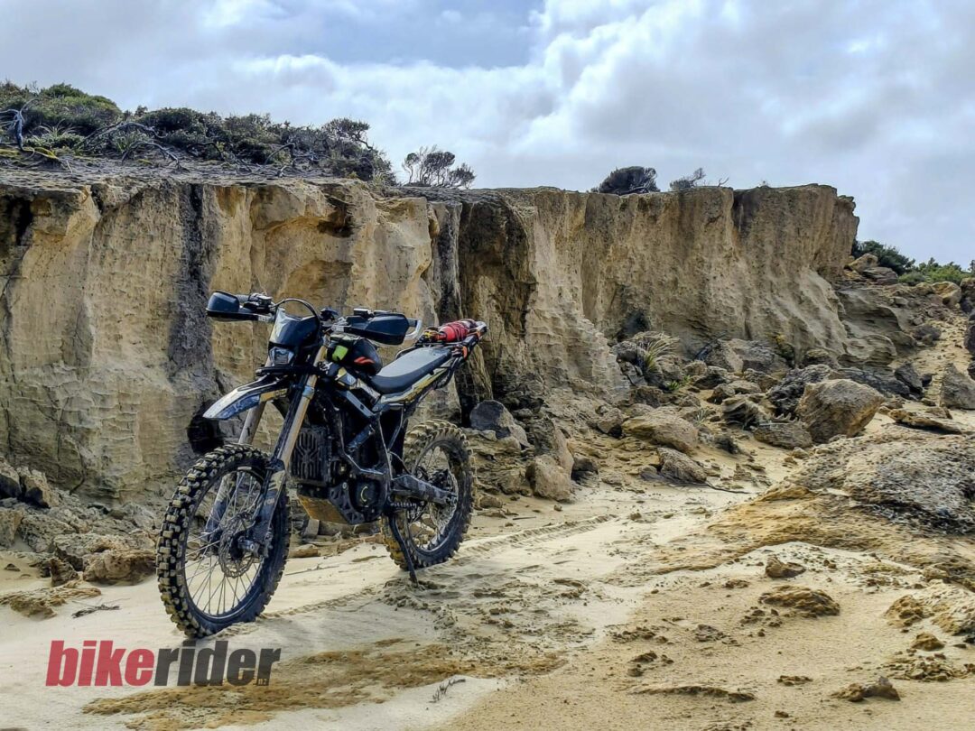Off-road motorcycle parked on a sandy beach beside eroded rock cliffs under a cloudy sky.