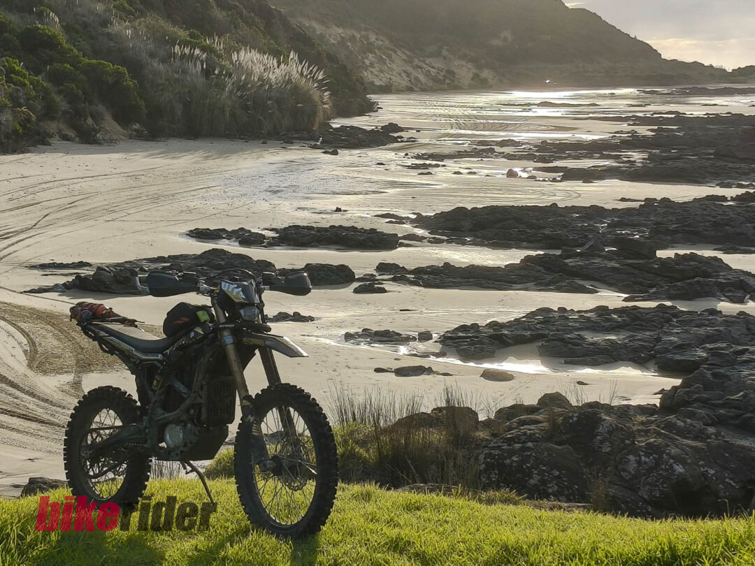 Adventure motorcycle parked on a sandy beach with rocks, shallow water, and grassy foreground; cliffs rise in the distance at dusk.