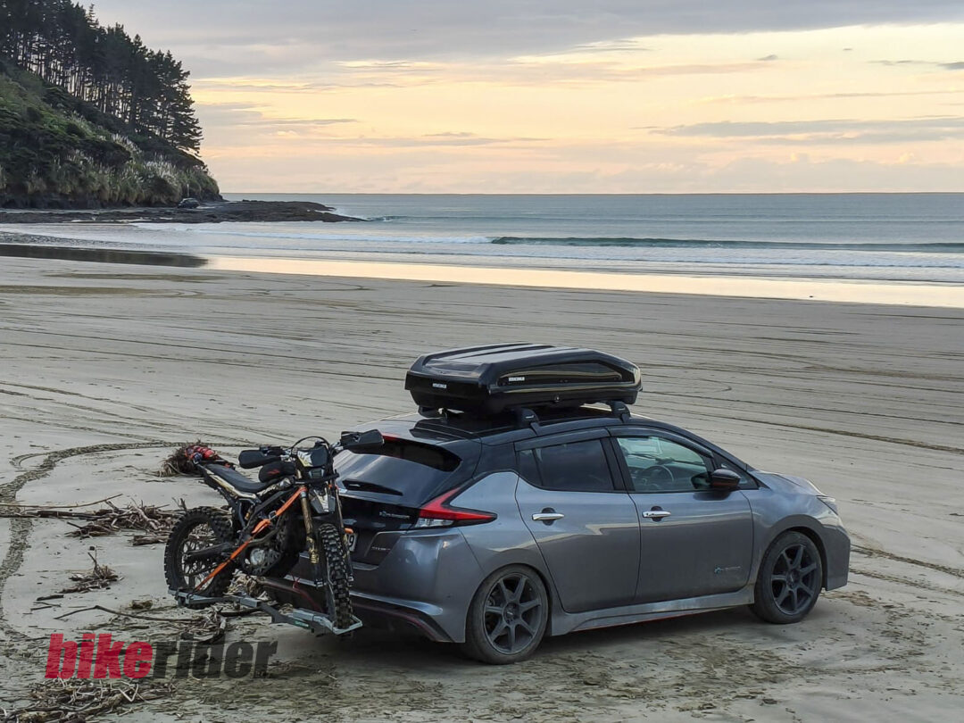 Car on a sandy beach with a roof-mounted bike rack holding two bikes, calm sea and sunset sky in the background.