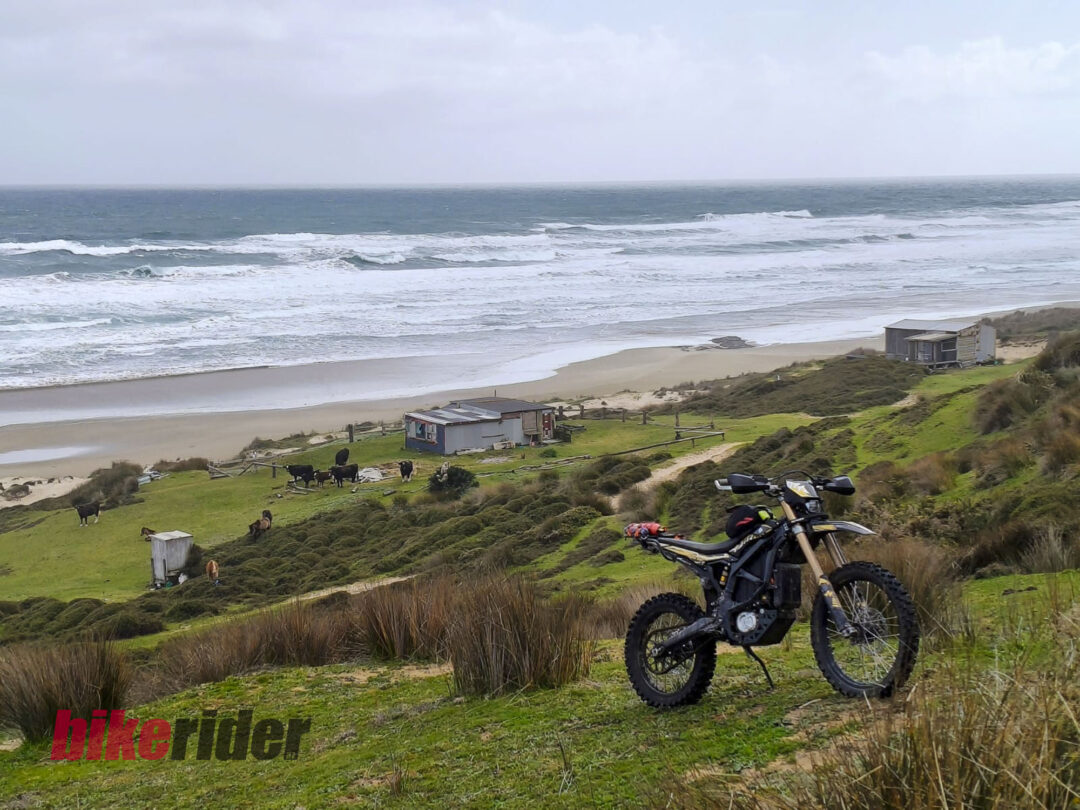 Coastal scene with waves crashing on a sandy beach, grassy dunes, small huts, grazing cows, and a dirt bike in the foreground.