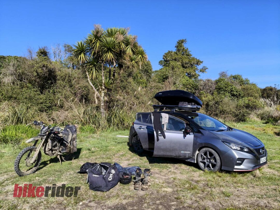 Silver SUV with a roof cargo box parked on a grassy area; dirt bikes and riding gear laid out in front amid shrubs and a clear blue sky.