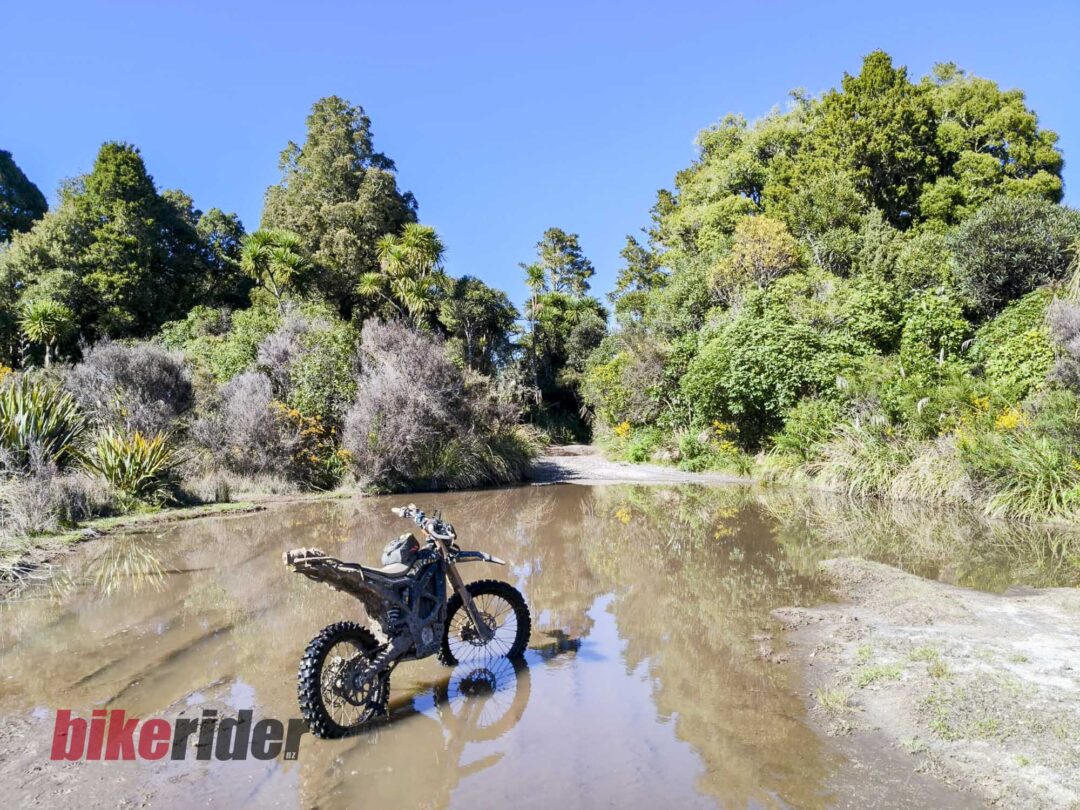 Off-road dirt bike splashing through a muddy pool with dense green trees and a clear blue sky behind.