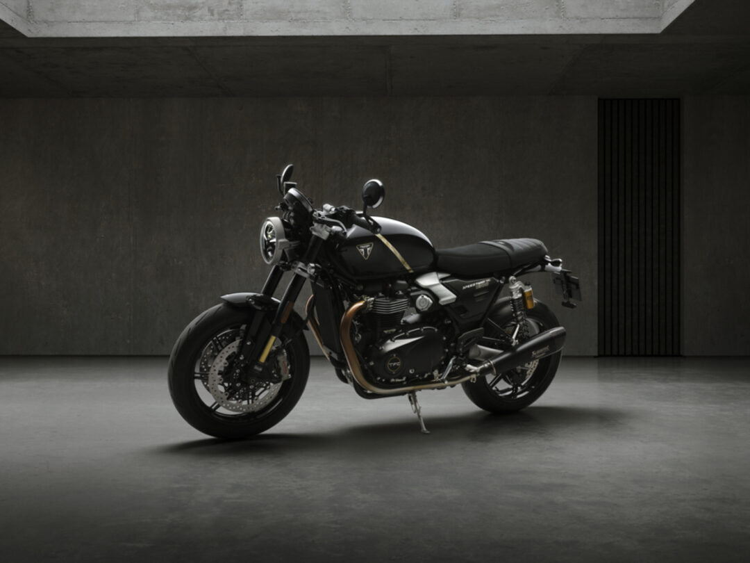 Black cafe-rac­er style motorcycle parked on a concrete garage floor, kickstand down, in front of a dark concrete wall with a vertical vent.
