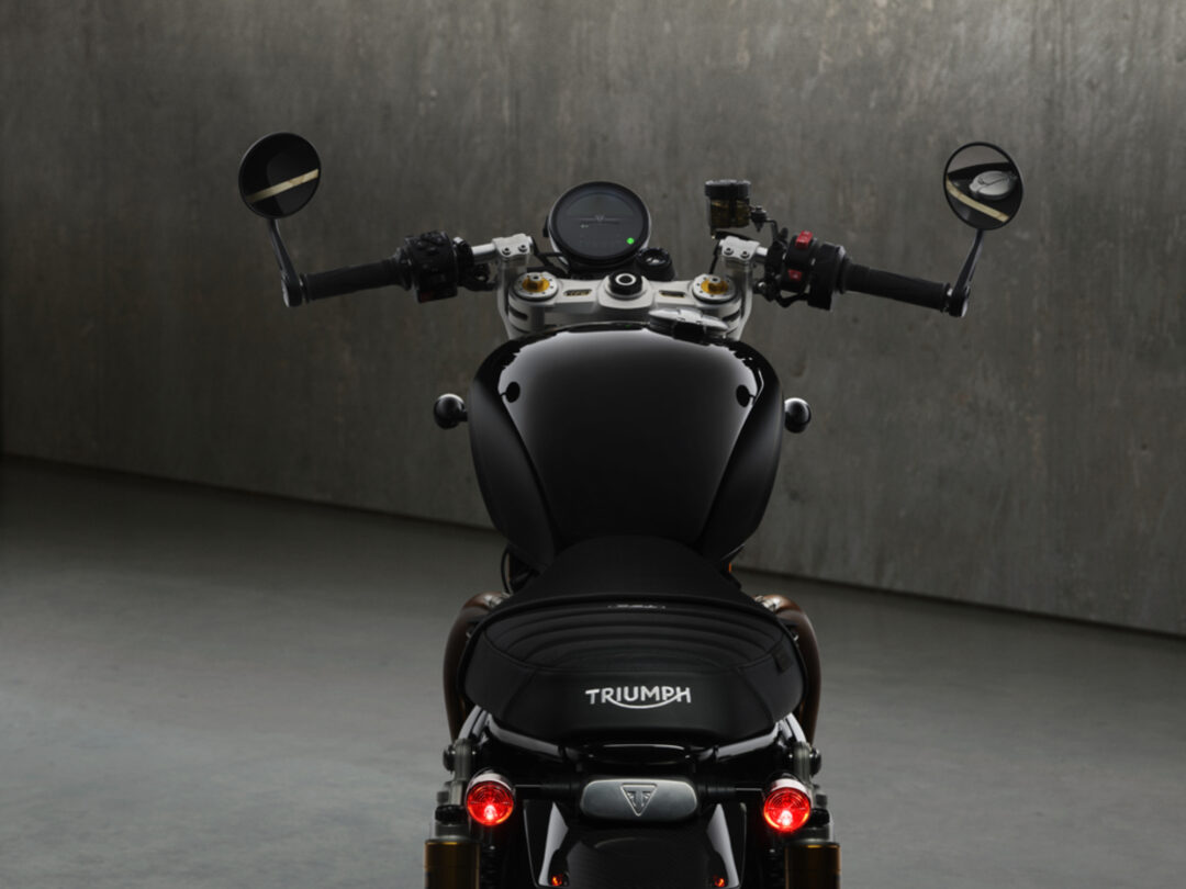 Rear view of a black Triumph motorcycle, handlebars and circular mirrors visible, in a dim backdrop with a glossy fuel tank and Triumph branding on the seat area.