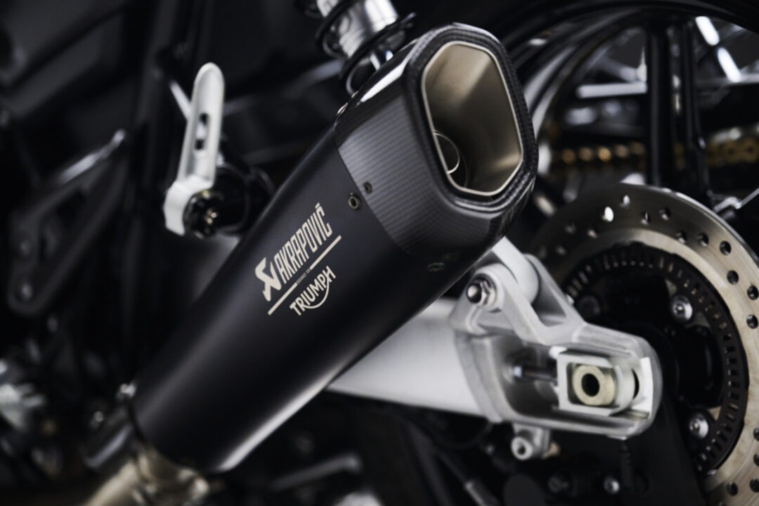 Close-up of a black Akrapovic exhaust on a Triumph motorcycle, focusing on the tip and branding logo, with surrounding mechanical parts visible