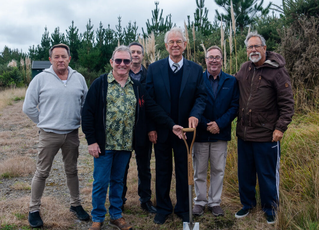 Six men stand together outdoors on a grassy, rocky area, one in a suit holding a shovel as if at a ceremonial start or groundbreaking.