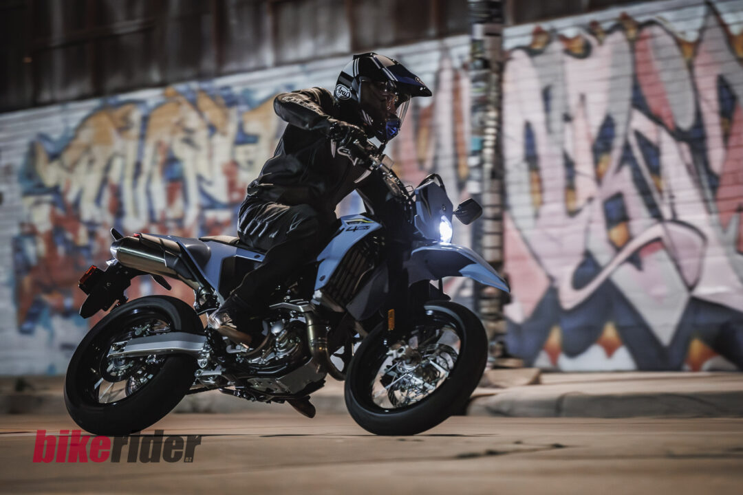 Motorcyclist in full gear performing a wheelie on a blue bike under graffiti-covered walls near an urban backdrop.