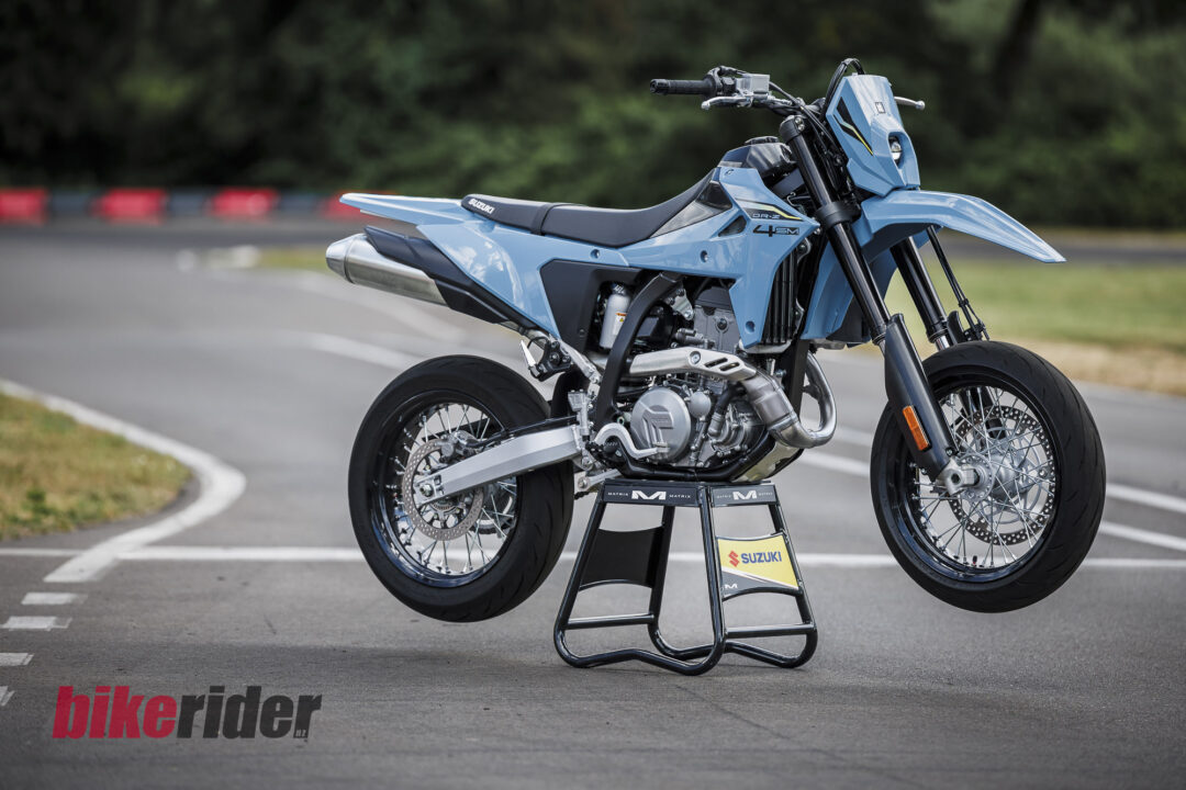 Blue Suzuki motorcycle on a stand outdoors, angled to show the side and front wheel near a race track backdrop.